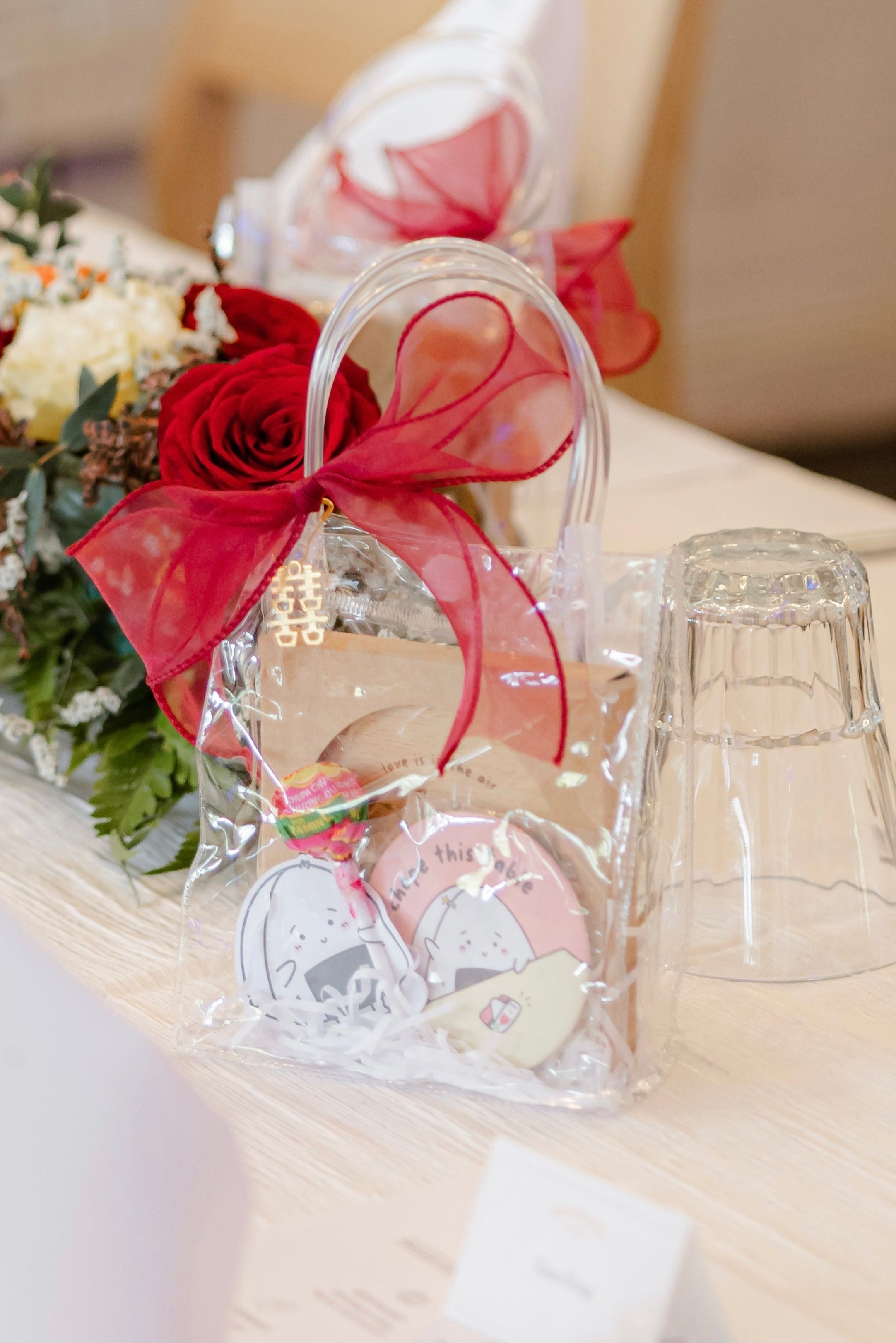 Decorative wedding table setting with wedding favour from Kooklesart, red roses, clear glass, and floral arrangements on a light surface.